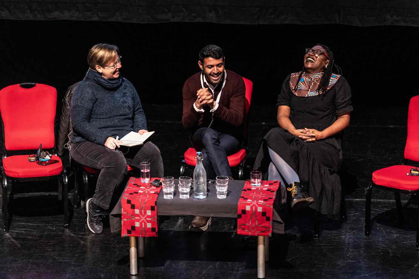 Three people seated on a stage on soft chairs and laughing, a small table in front of them with cups and bottles filled with water.