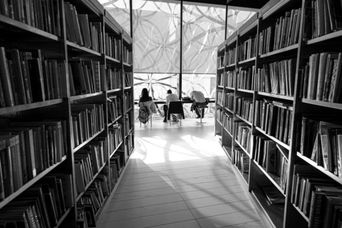 Two rows of books and three people working at desks at the end, in front of a window.